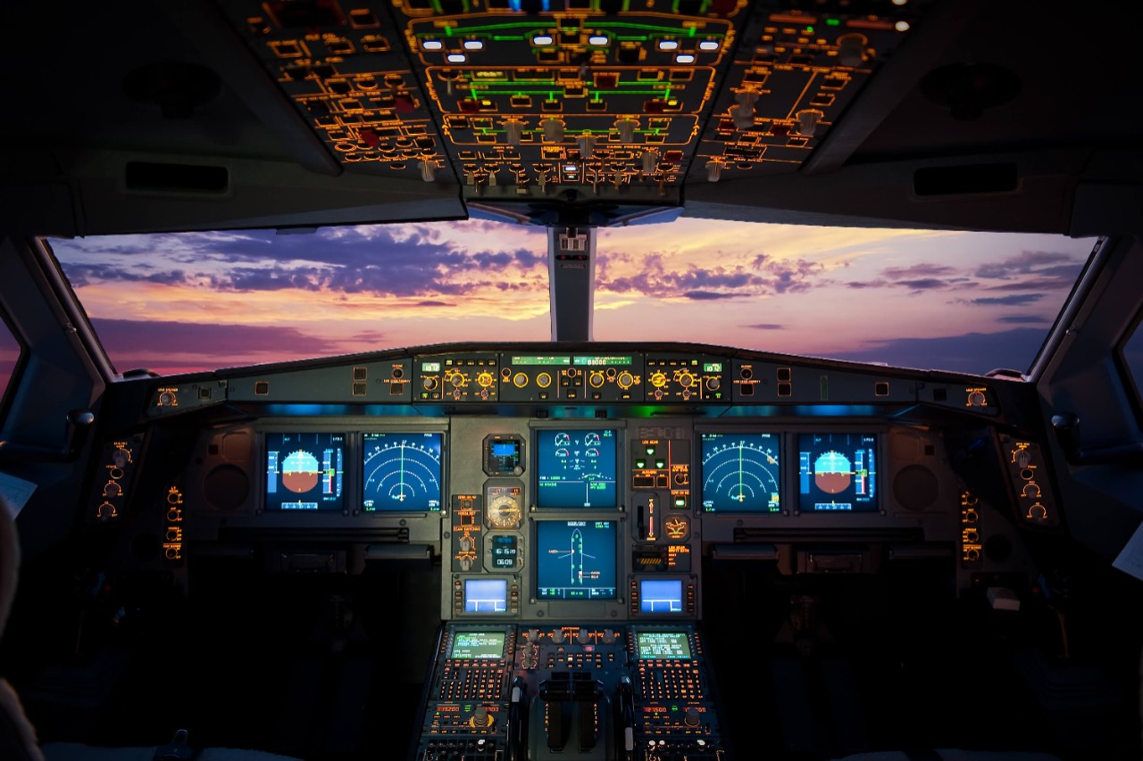 An illuminated commercial airliner cockpit looking out over an evening sunset sky
