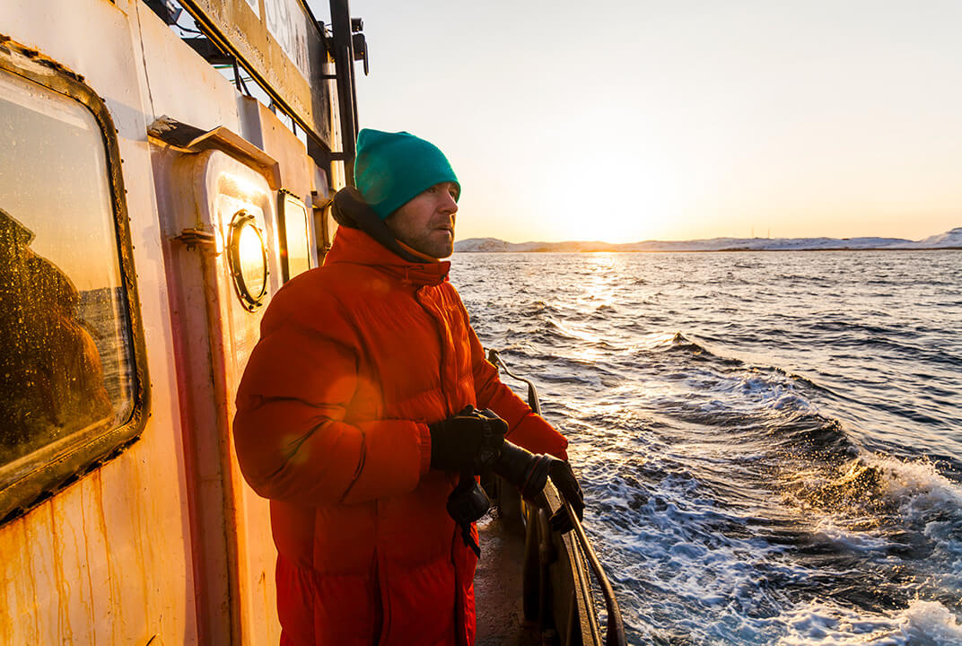 Sailor wrapped up in hat and coat standing on deck in a rough sea as the sun sets