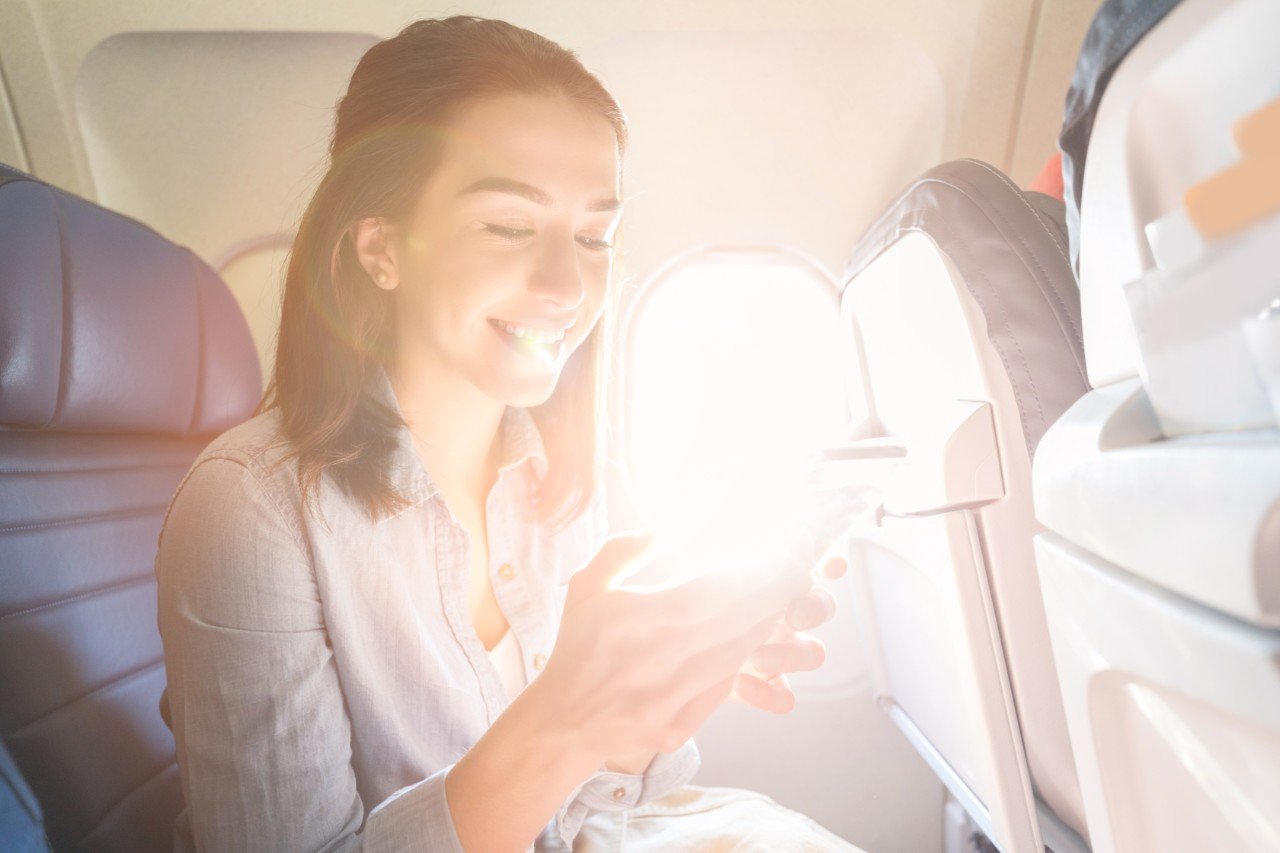 Cheerful young woman uses smart phone while on a commercial flight. The sun is streaming through the window next to her.