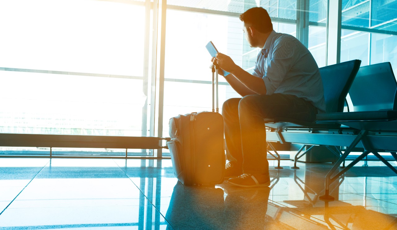Traveller sits in at chair at an airport terminal waiting for a plane. He holds a digital tablet and looks out through the windows towards the planes. The sun shines. A suitcase stands infront of the man.