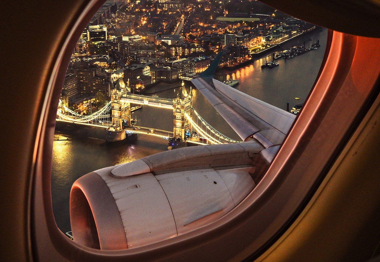 Looking out across a wing and engine on a commercial airliner flying over London's Tower Bridge which is illuminated