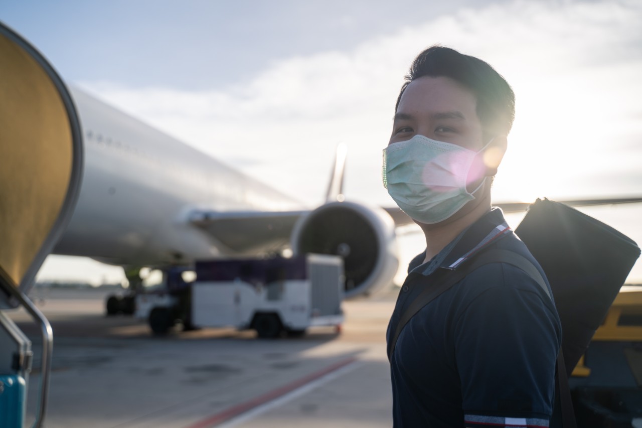 Asian man wearing face mask walks to stair entering airplane, parking at remote bay located outside terminal in airport. Male passenger traveling by plane transportation during covid19 virus pandemic.