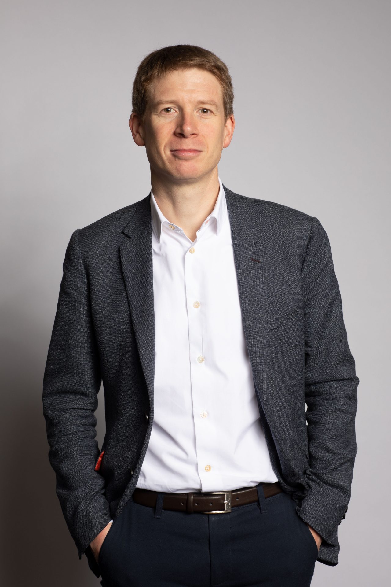 man stands with hands in pockets wearing a grey suit and white shirt