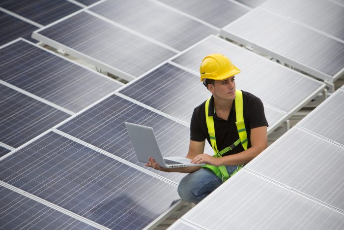 Man in hardhat next to solar panels using laptop