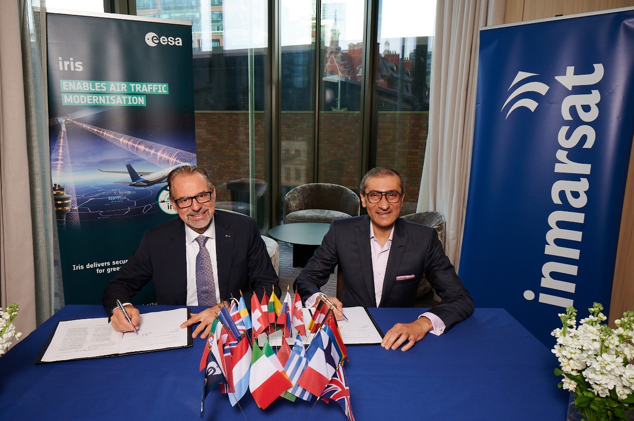 two men sit at a table with flags signing  the new contract