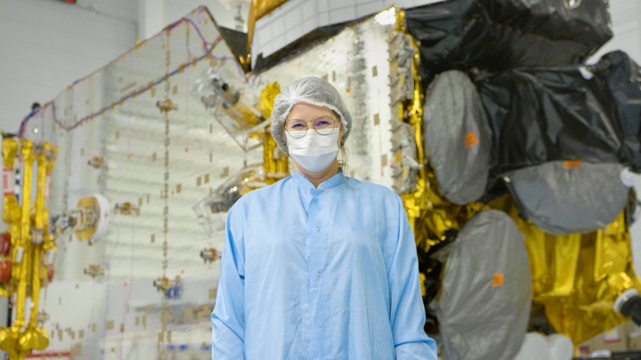 Kristiina Jokela wears a lab coat, hairnet and mask as she stands in front of the I-6 F1 satellite in the testing facility