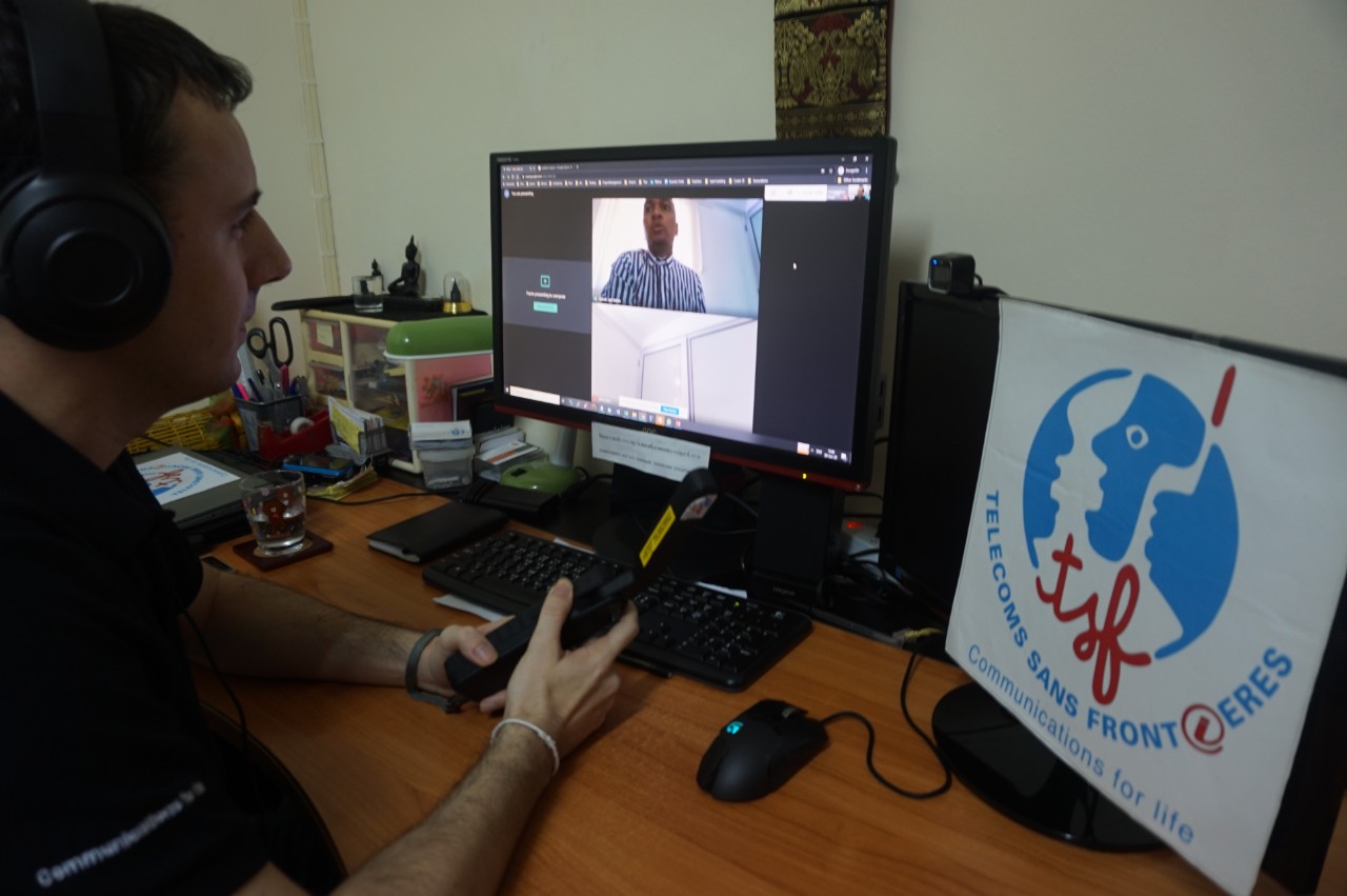 A TSF technician sitting at a desk in front of his computer remotely training an official in Mozambique on how to use and Inmarsat's IsatPhone 2 handheld satellite phone