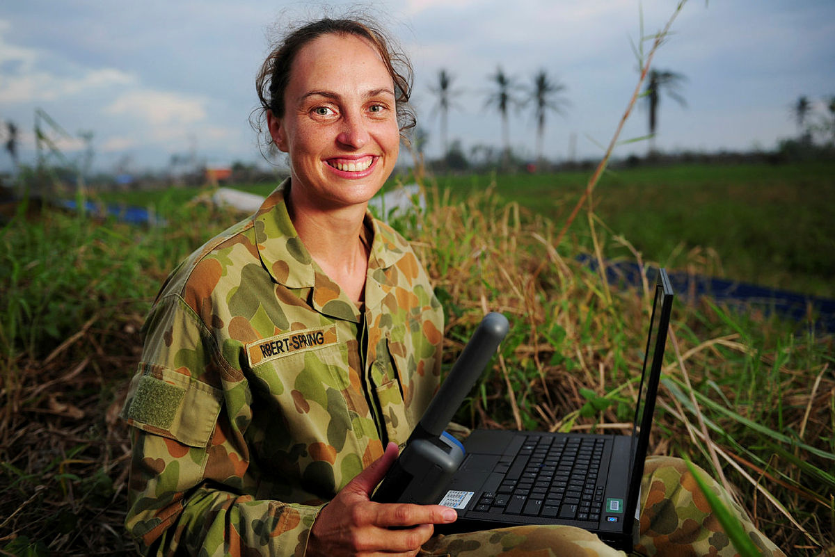 Female Australian Army Signaller sitting outside with a laptop and holding an Inmarsat IsatPhone handheld satellite phone