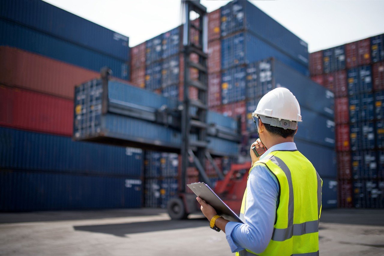 Man with clipboard examining freight containers with a fork lift working in the background 