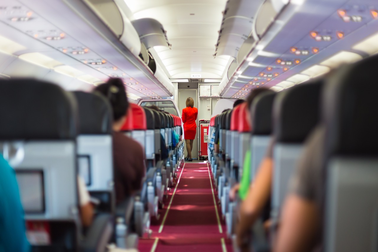 Interior of airplane with passengers on seats and stewardess in red uniform at front of the aisle